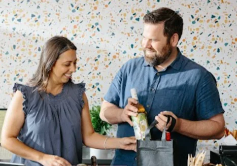 a man and a woman both wearing blue, pack a bag and laugh together.